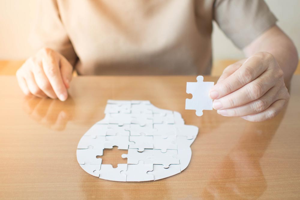 Elderly woman hands holding missing white jigsaw puzzle piece down into the place as a human head brain shape. Creative idea for memory loss, dementia, Alzheimer's disease and mental health concept